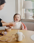 Woman feeds smiling teething baby in high chair with Mushie First Feeding Baby Spoons by window.