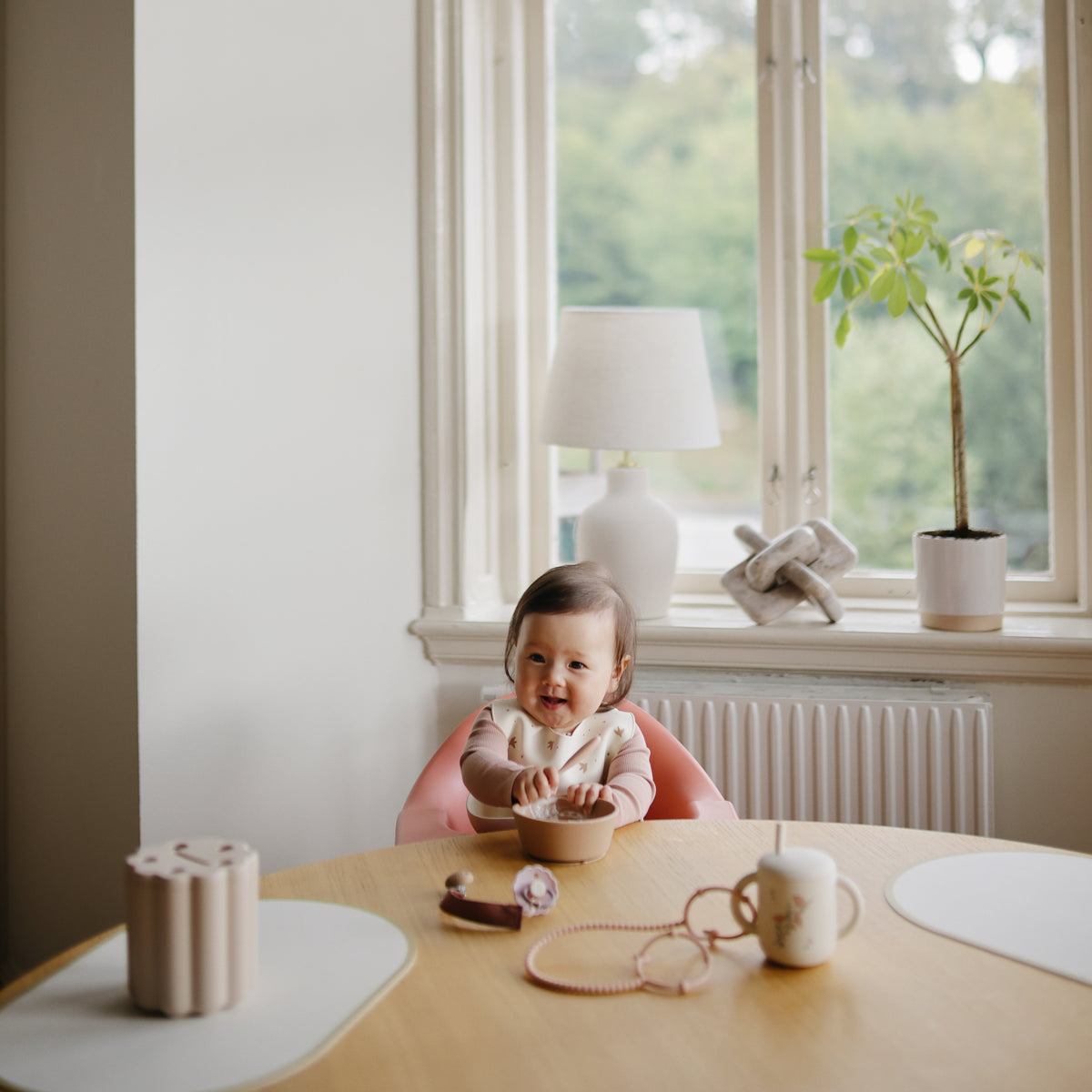 Baby in high chair with mushie Linen Pacifier Clip, eating at a table in a bright room.