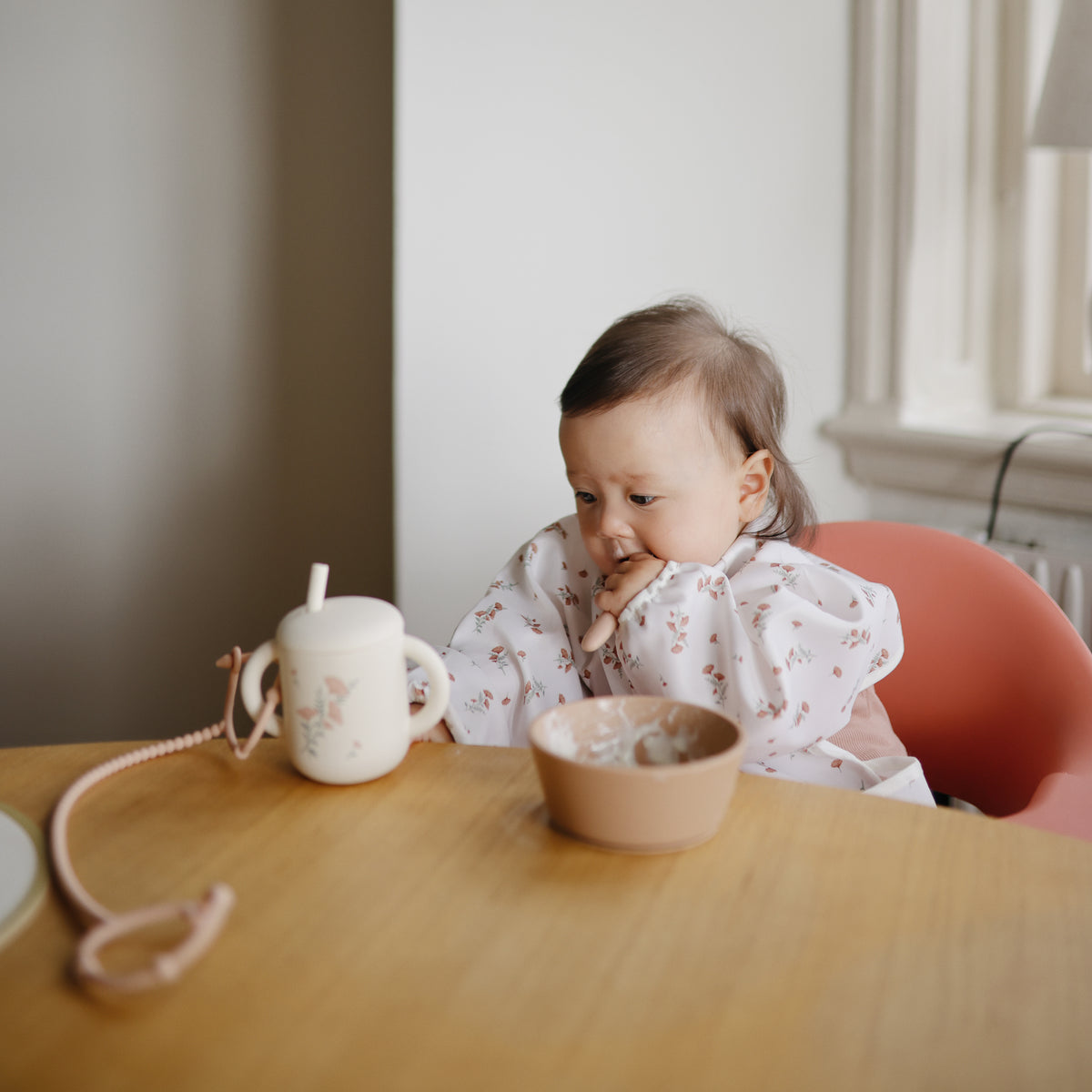 Baby wearing a mushie Long Sleeve Bib in a high chair with a bowl and cup.