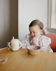 Baby wearing a mushie Long Sleeve Bib in a high chair with a bowl and cup.