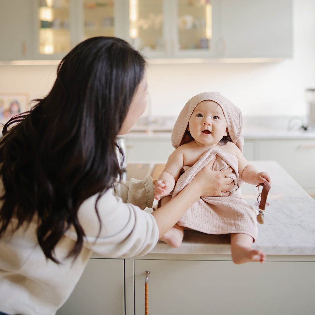 Smiling baby with mushie Linen Pacifier Clip 2-Pack on kitchen counter, held by mom.