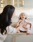 Smiling baby with mushie Linen Pacifier Clip 2-Pack on kitchen counter, held by mom.