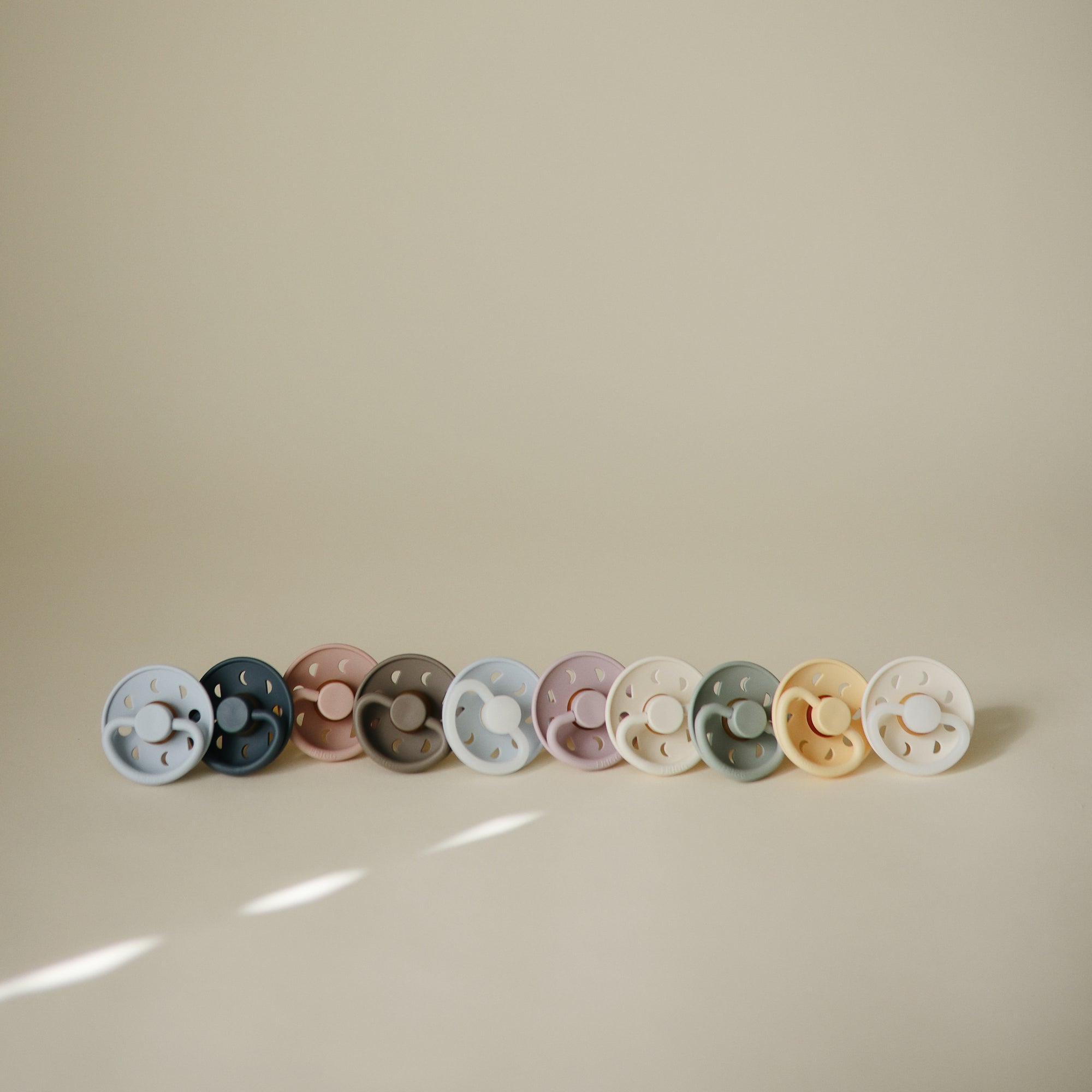 Nine FRIGG Moon Natural Rubber Pacifier 2-Packs lined up on a neutral background.