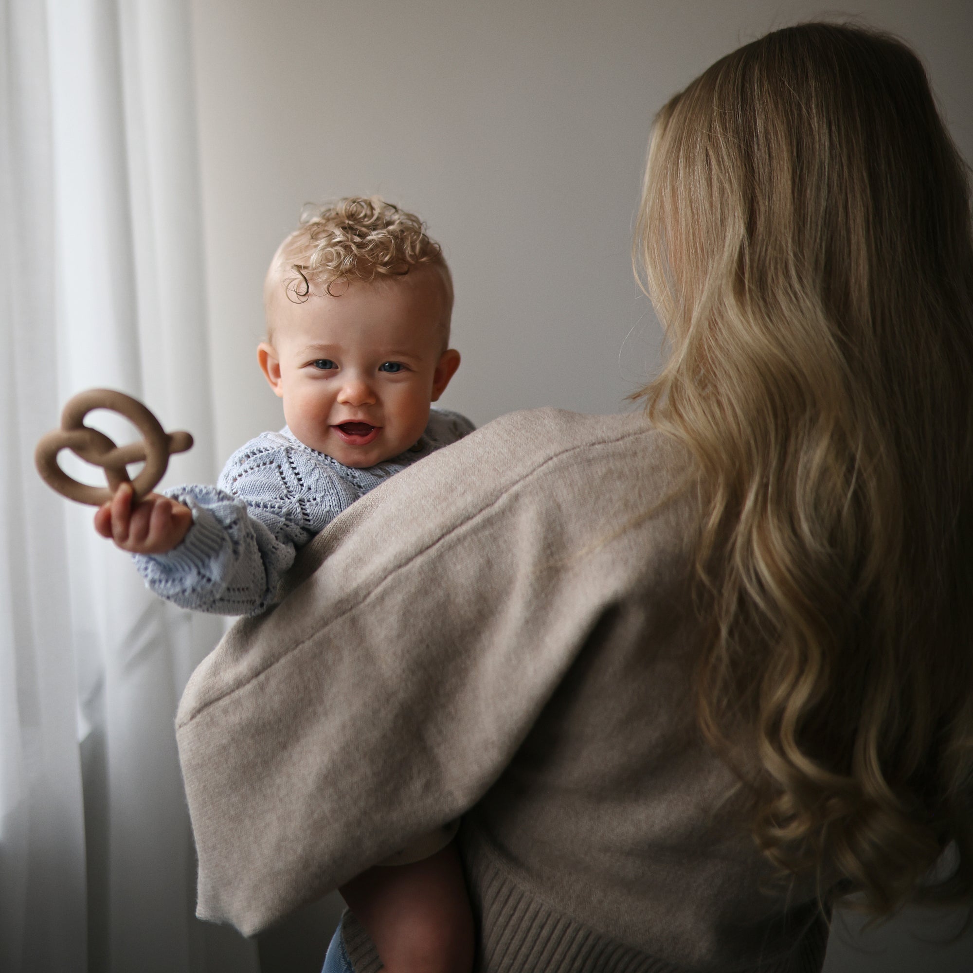 A baby with a mushie Pretzel Teether smiles while held by a woman with long blonde hair.