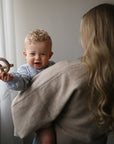 A baby with a mushie Pretzel Teether smiles while held by a woman with long blonde hair.