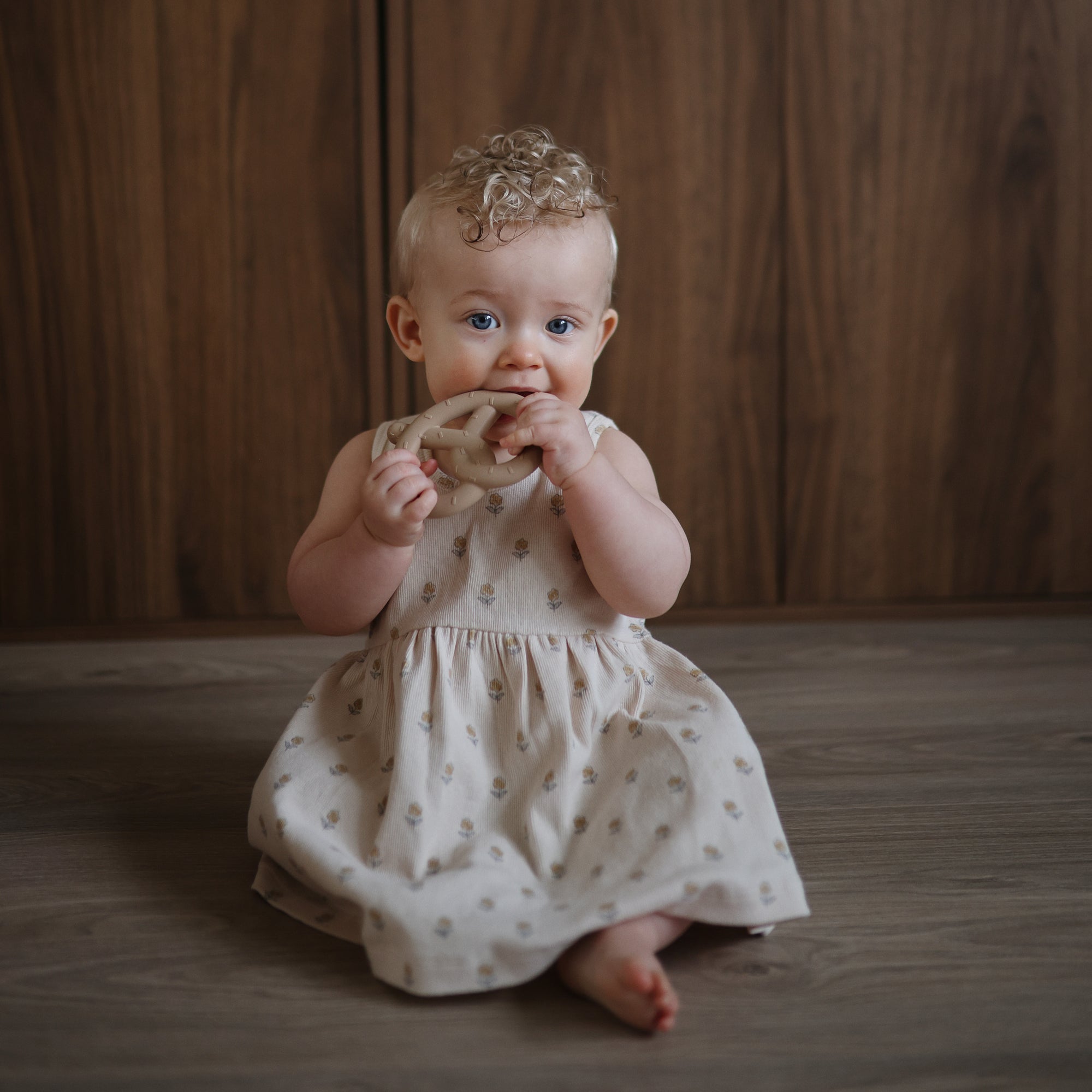 A baby in a beige dress smiles on a wooden floor, holding the mushie Pretzel Teether.