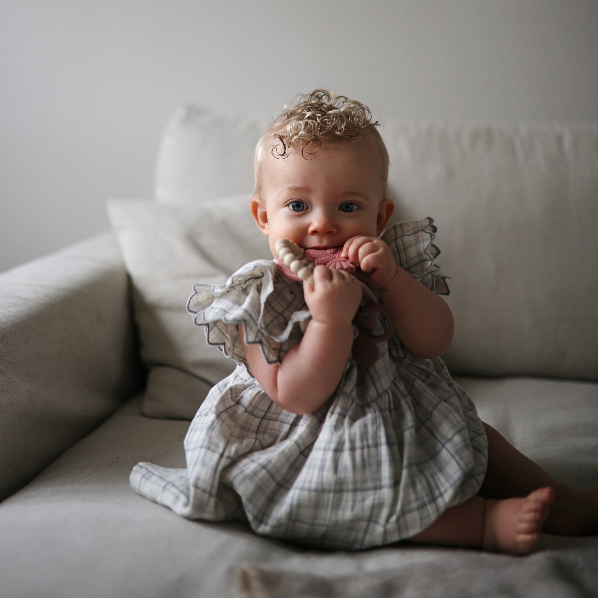 Baby in a plaid dress smiles on a couch, holding a mushie Teething Ring.