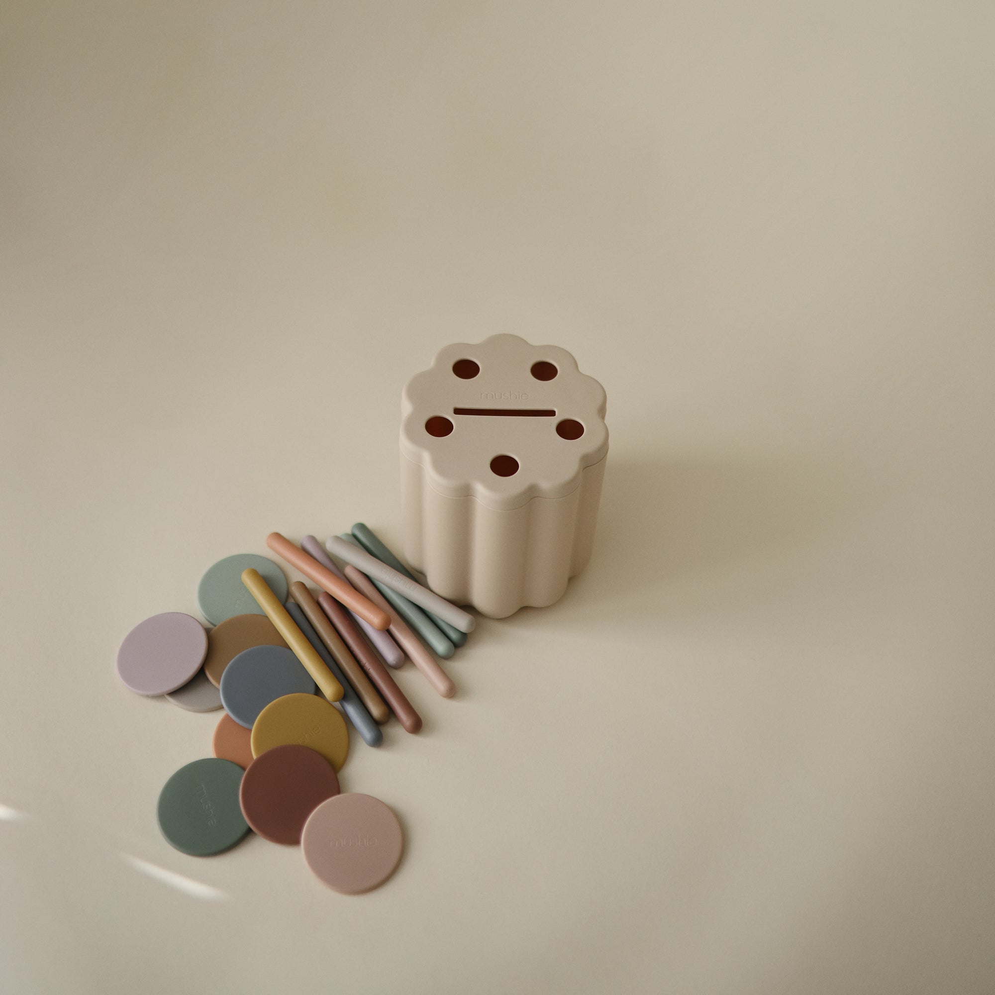 The mushie Coin &amp; Tube Sorting Set, a beige, flower-shaped educational toy with five holes, sits on a light surface surrounded by pastel disks and sticks that help children develop fine motor skills through play.