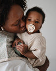 A woman lovingly kisses a baby on the cheek. The baby, wearing beige and holding a rattle, looks at the camera with a FRIGG Andersen Fairytale Silicone Pacifier from FRIGG in their mouth. The background is softly blurred.