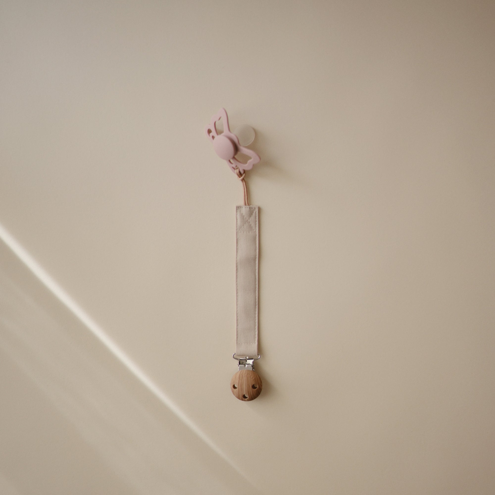 A pink butterfly-shaped pacifier clip hangs on a beige wall, paired with the FRIGG Butterfly Anatomical Silicone Pacifier 2-Pack (6-18 Months). The light strap features a wooden clip, combining charm and comfort from FRIGG for your baby.