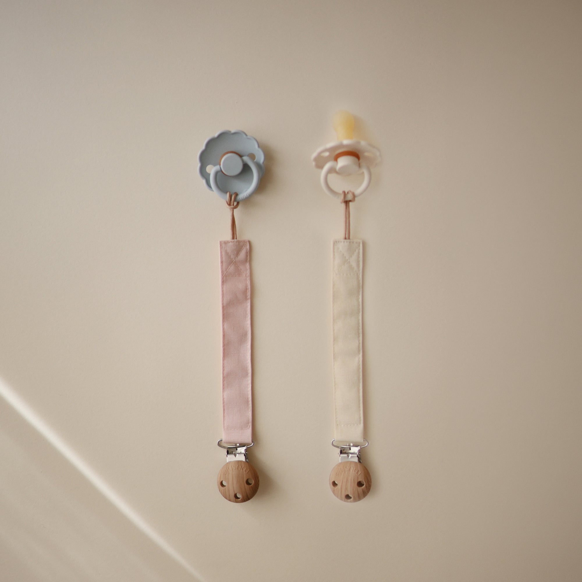 Two mushie Linen Pacifier Clips (2-Pack) with silicone clasps hang on a beige wall.
