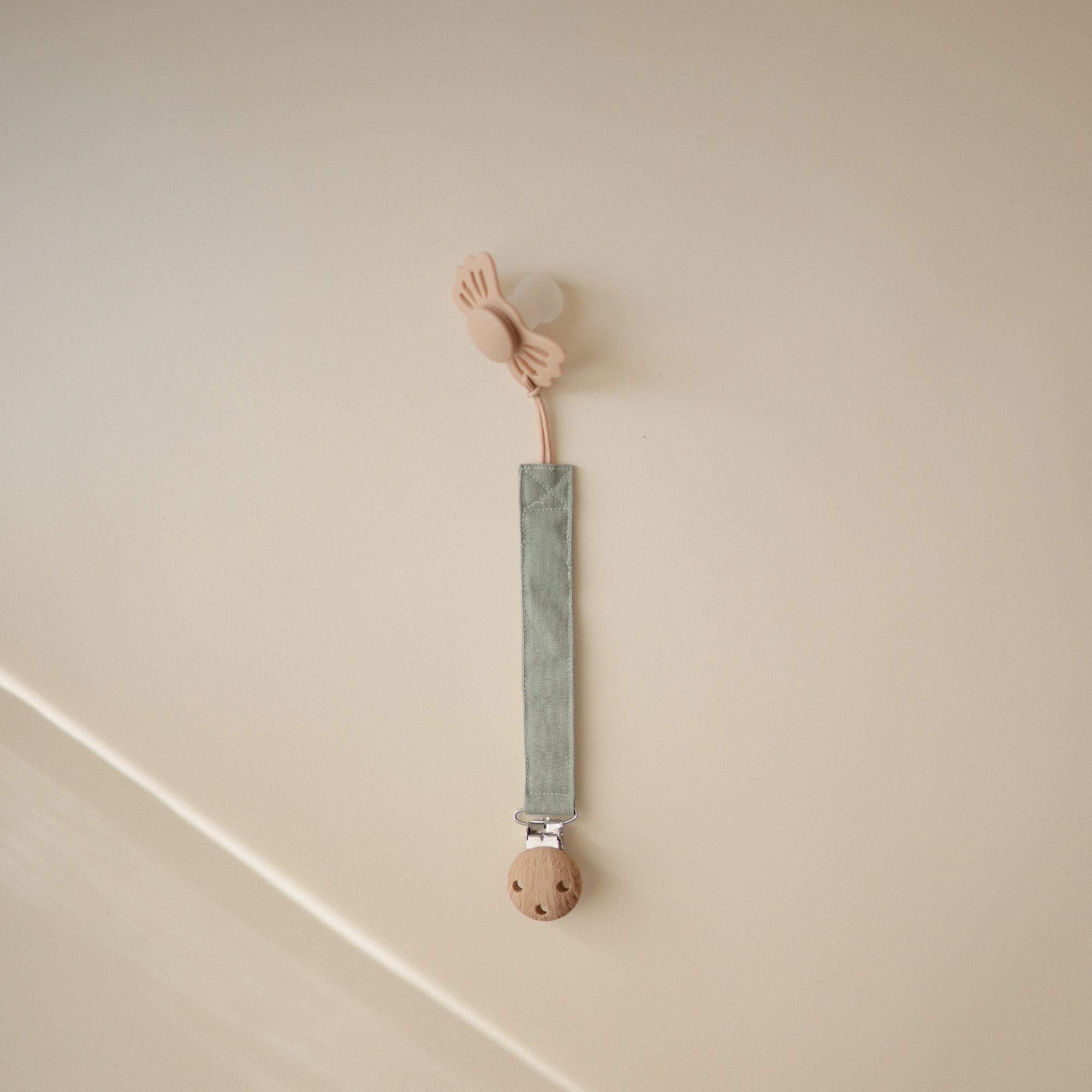 The FRIGG Lucky Symmetrical Silicone Pacifier (0-6 Months) is shown in beige, attached to a sage green fabric clip with a wooden clasp, hanging against a light beige wall.