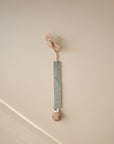 The FRIGG Lucky Symmetrical Silicone Pacifier (0-6 Months) is shown in beige, attached to a sage green fabric clip with a wooden clasp, hanging against a light beige wall.