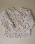 Mushie Long Sleeve Bib in a red floral print with a front pocket and wipe-clean fabric.