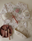 Mushie Long Sleeve Bib, silicone bib, divided plate, two cups, two spoons, and a teething toy.