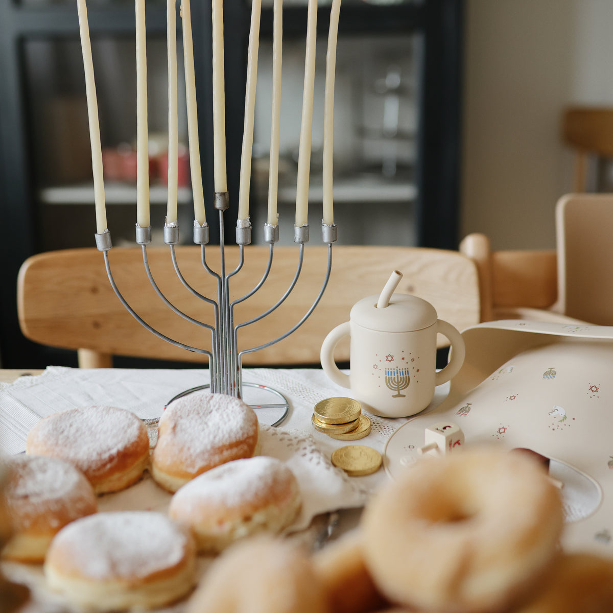 A menorah, donuts, chocolate coins, and a Mushie Silicone Training Cup + Straw on a Hanukkah table.