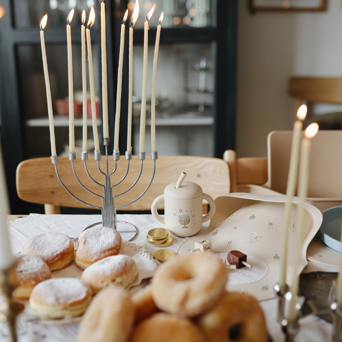 A lit menorah, donuts, and gelt sit by a mushie Silicone Baby Bib for Hanukkah fun.