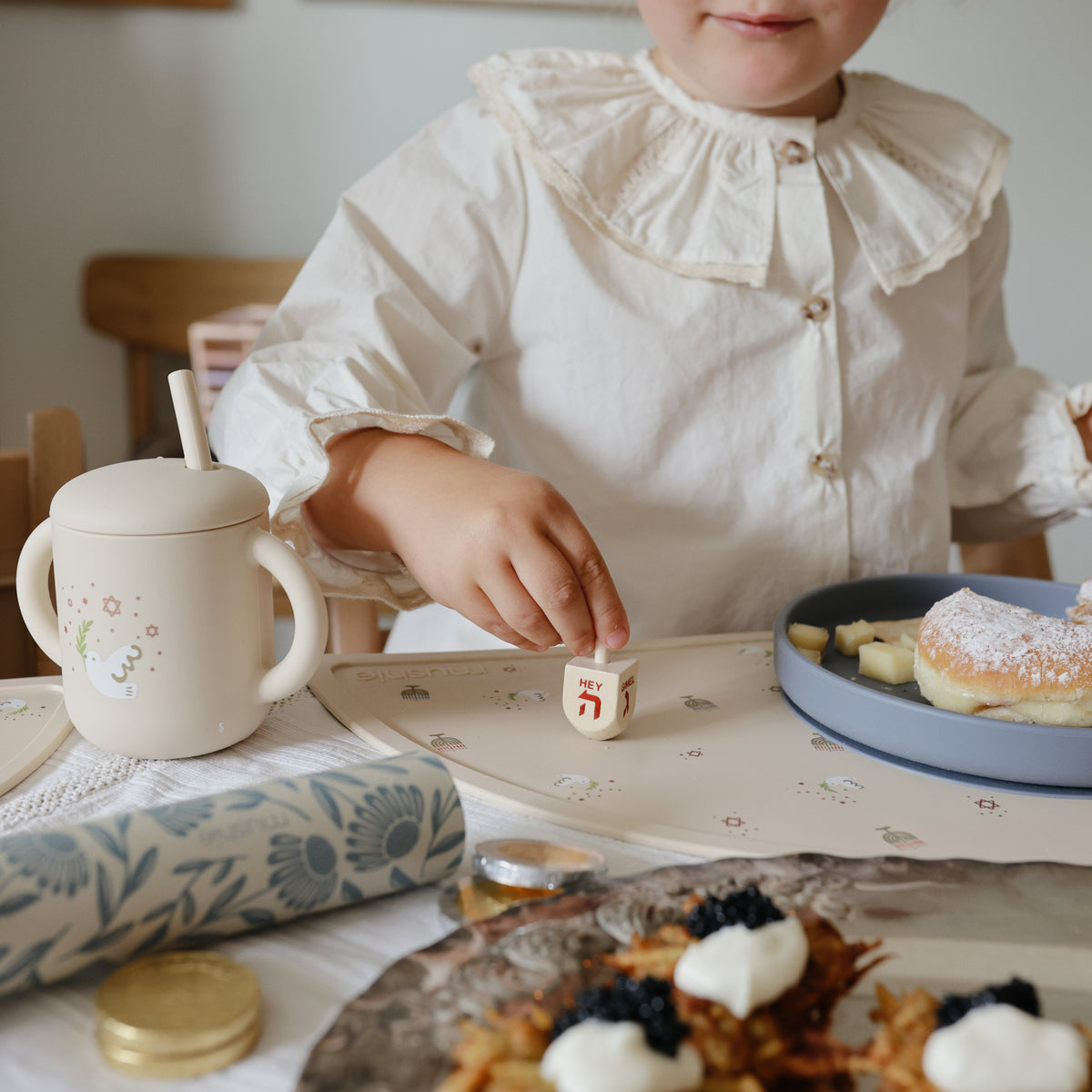 Child plays dreidel by sufganiyot, latkes, and Mushie Silicone Training Cup + Straw.