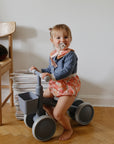 Toddler with pacifier sits on a mushie Mini Toddler Bike indoors next to a stack of magazines.