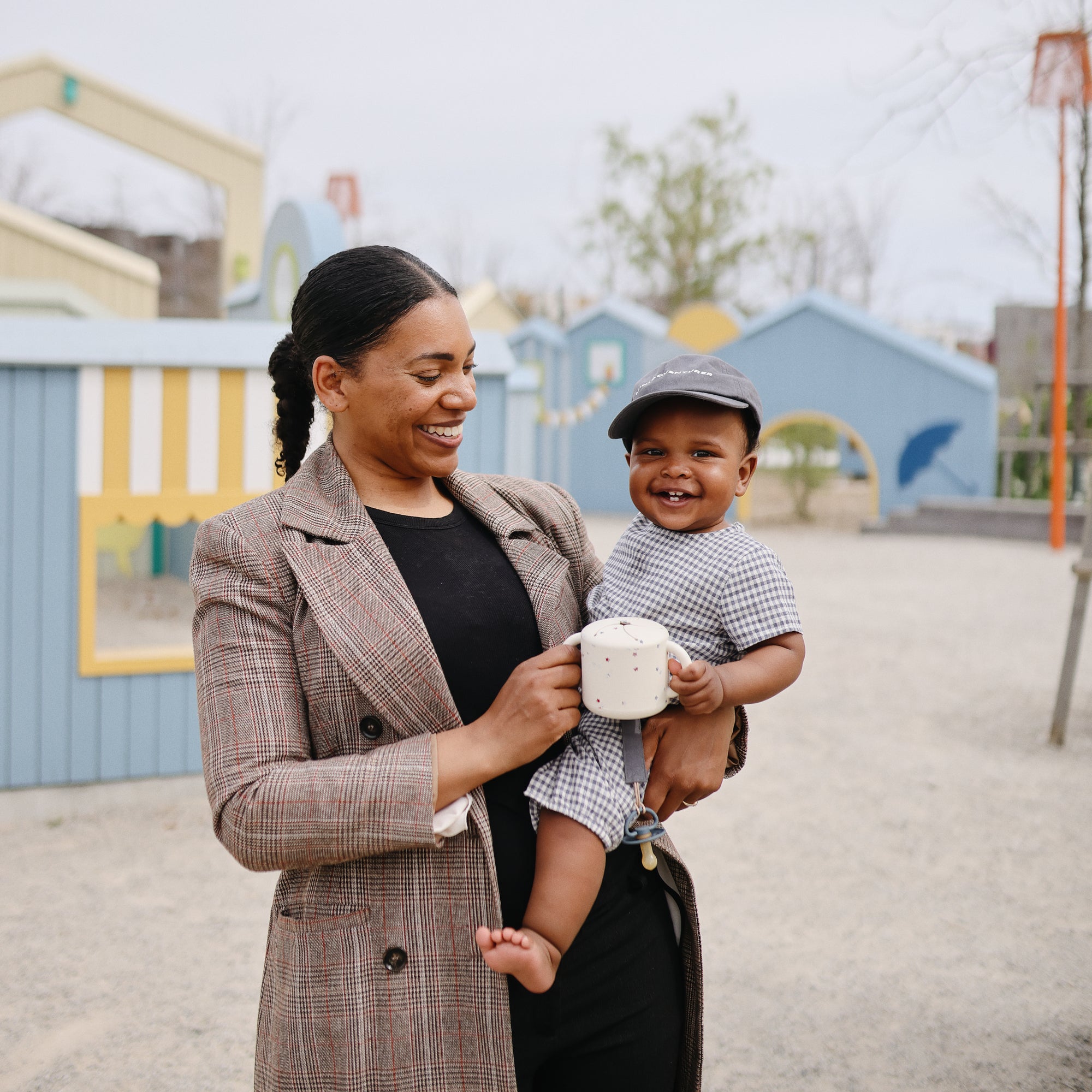Smiling woman holding a baby and a mushie Snack Cup at an outdoor playground.