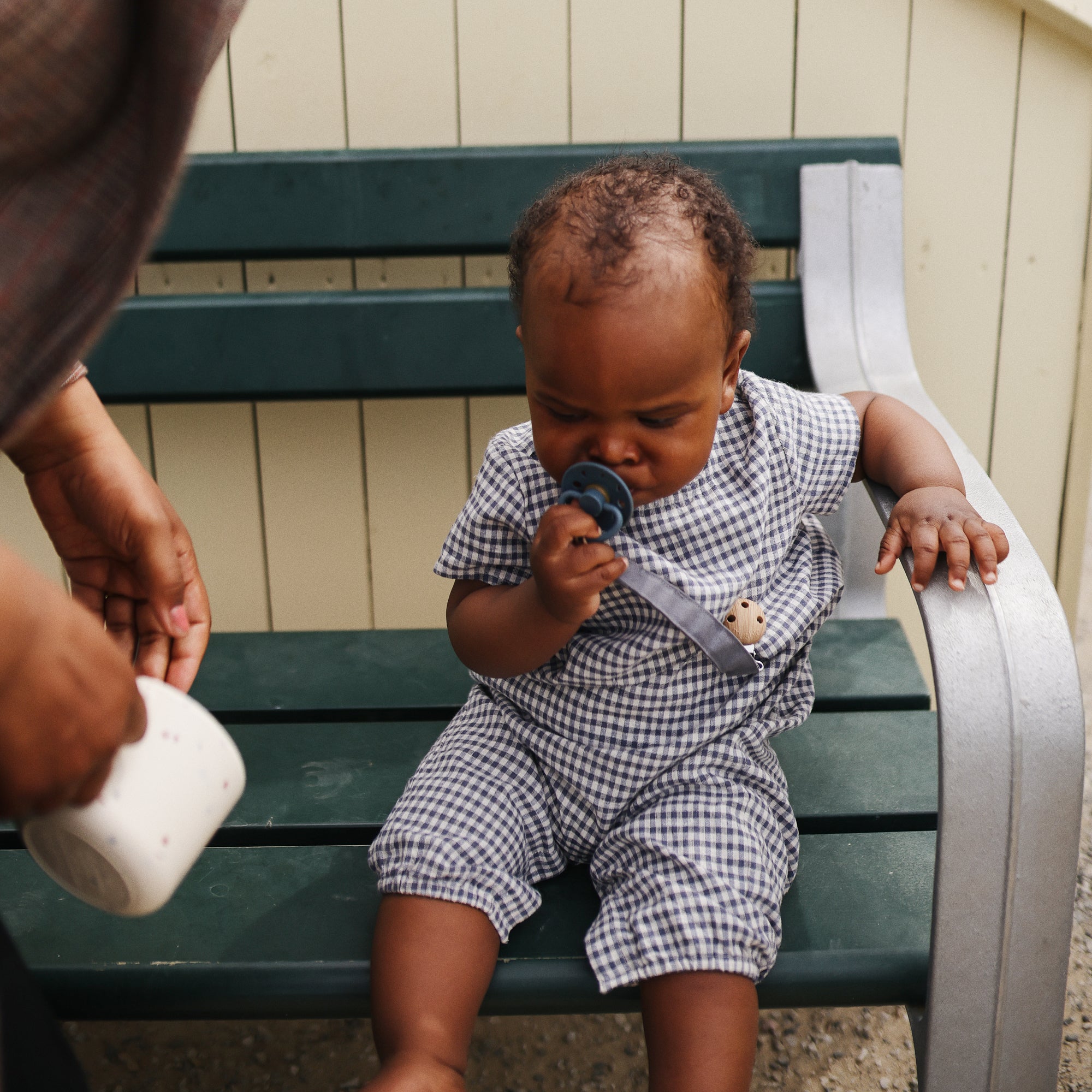 Baby in checkered outfit holds FRIGG Moon Natural Rubber Pacifier on green bench with adult.
