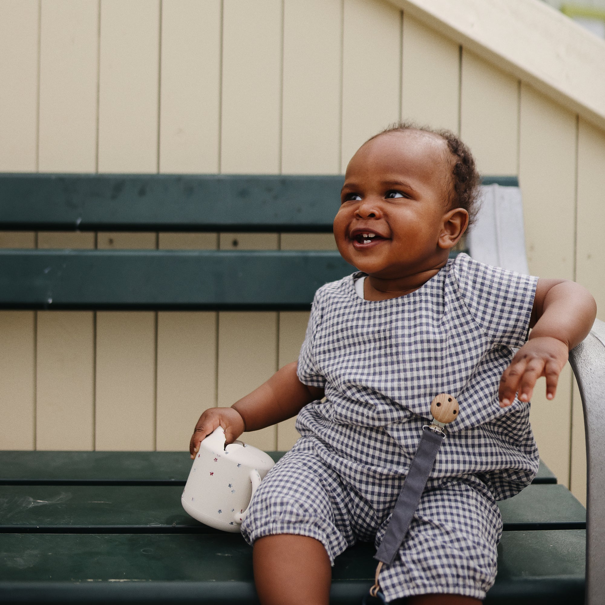 Smiling baby in checkered outfit sits on bench holding mushie Linen Pacifier Clip 2-Pack.