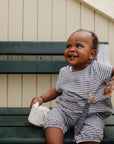 Smiling baby in checkered outfit sits on bench holding mushie Linen Pacifier Clip 2-Pack.