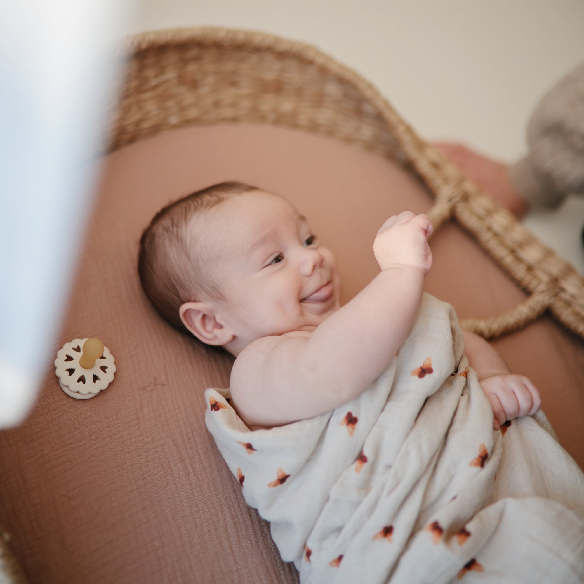 A baby smiles in a woven bassinet wrapped in a light-patterned blanket, while a FRIGG Andersen Fairytale Natural Rubber Pacifier 2-Pack rests nearby on the mattress.