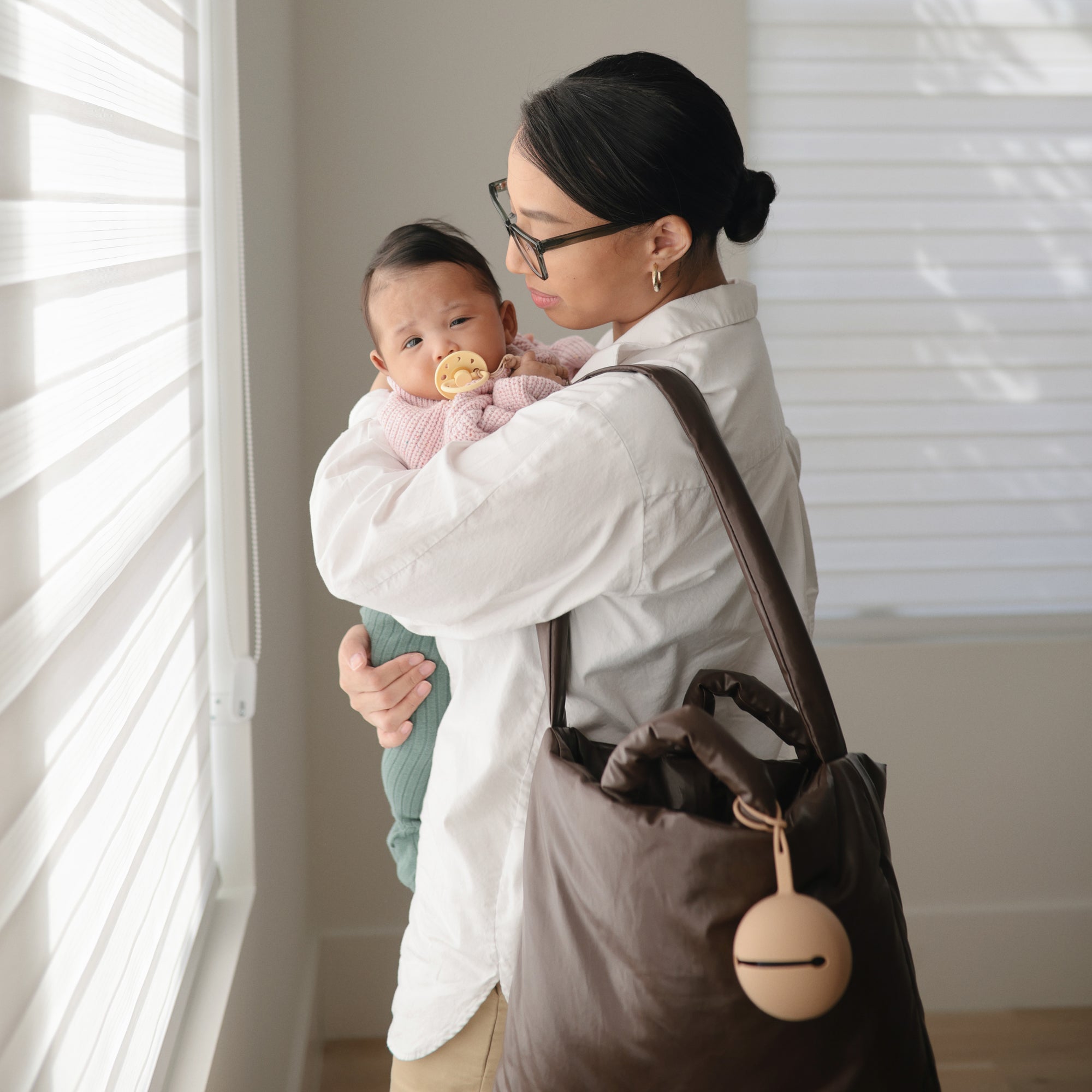 A woman in glasses and a white shirt holds a baby with the FRIGG Moon Natural Rubber Pacifier 2-Pack by FRIGG. She carries a large brown bag with baby essentials and a round keychain, standing by a window with blinds in soft light.