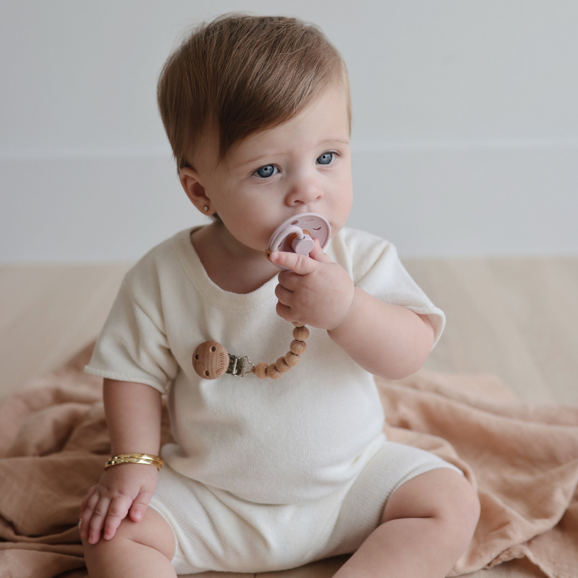 A baby with light brown hair and blue eyes, dressed in cream, sits on a beige blanket holding a FRIGG Moon Silicone Pacifier—one of the must-have baby essentials. A gold bracelet adorns their wrist as they look slightly to the side.