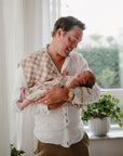 A man in a white shirt smiles as he gently holds a newborn wrapped in the mushie Stretchy Swaddle near a window with green plants on the sill, both bathed in soft natural light.