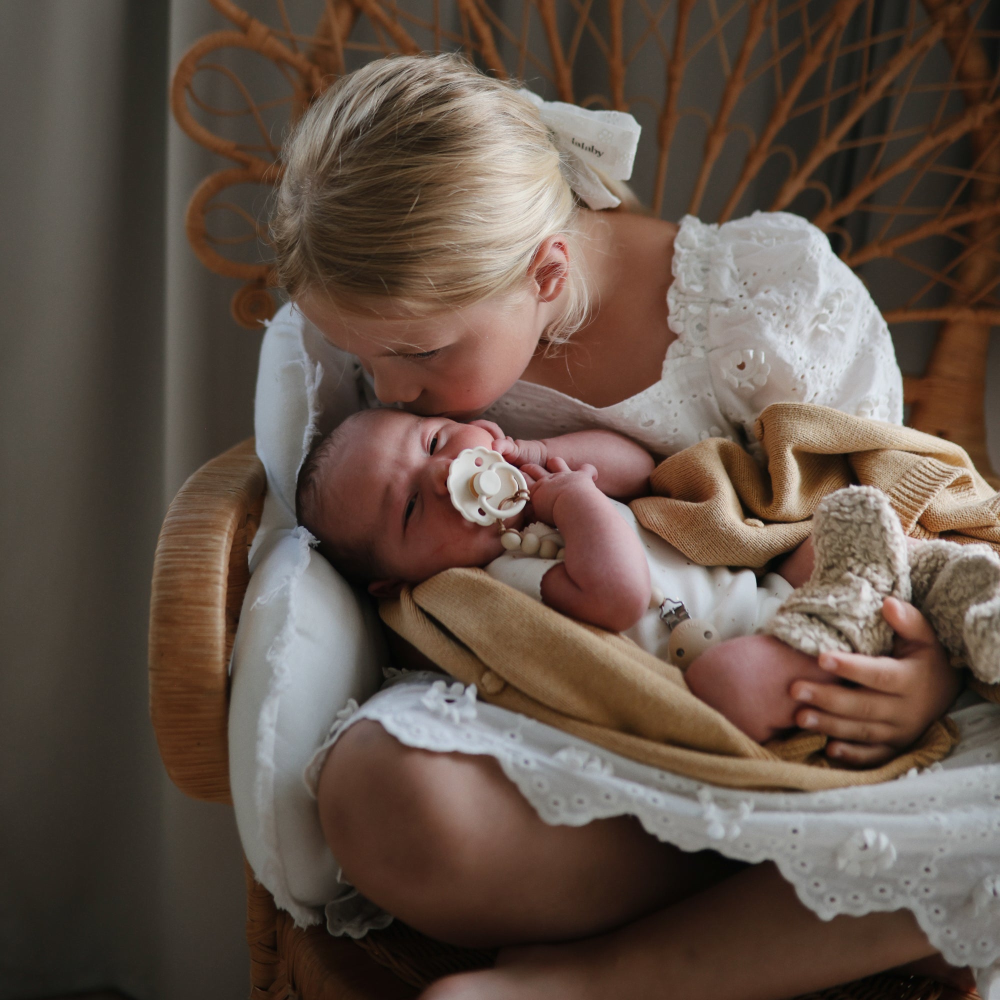 A young girl kisses a swaddled baby holding a FRIGG Daisy Natural Rubber Pacifier.