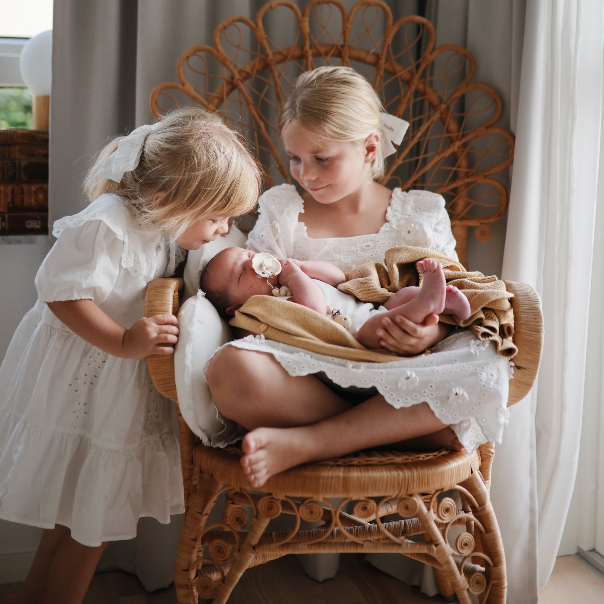 Two girls admire a newborn with a FRIGG Daisy Natural Rubber Pacifier in a cozy wicker chair.