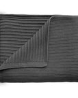 The mushie Knitted Ribbed Baby Blanket in dark gray is neatly folded with a corner turned over, showing its textured ribbed design on both sides against a white background.