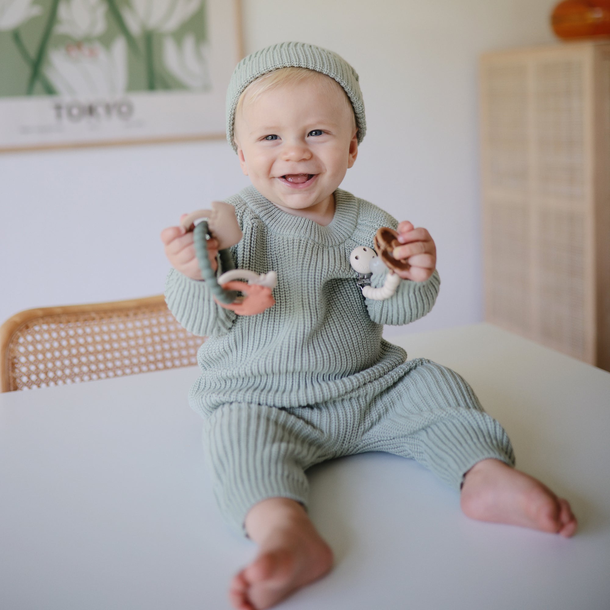 Smiling baby in green knit outfit holds mushie Teething Ring on a table.