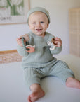 Smiling baby in green knit outfit holds mushie Teething Ring on a table.