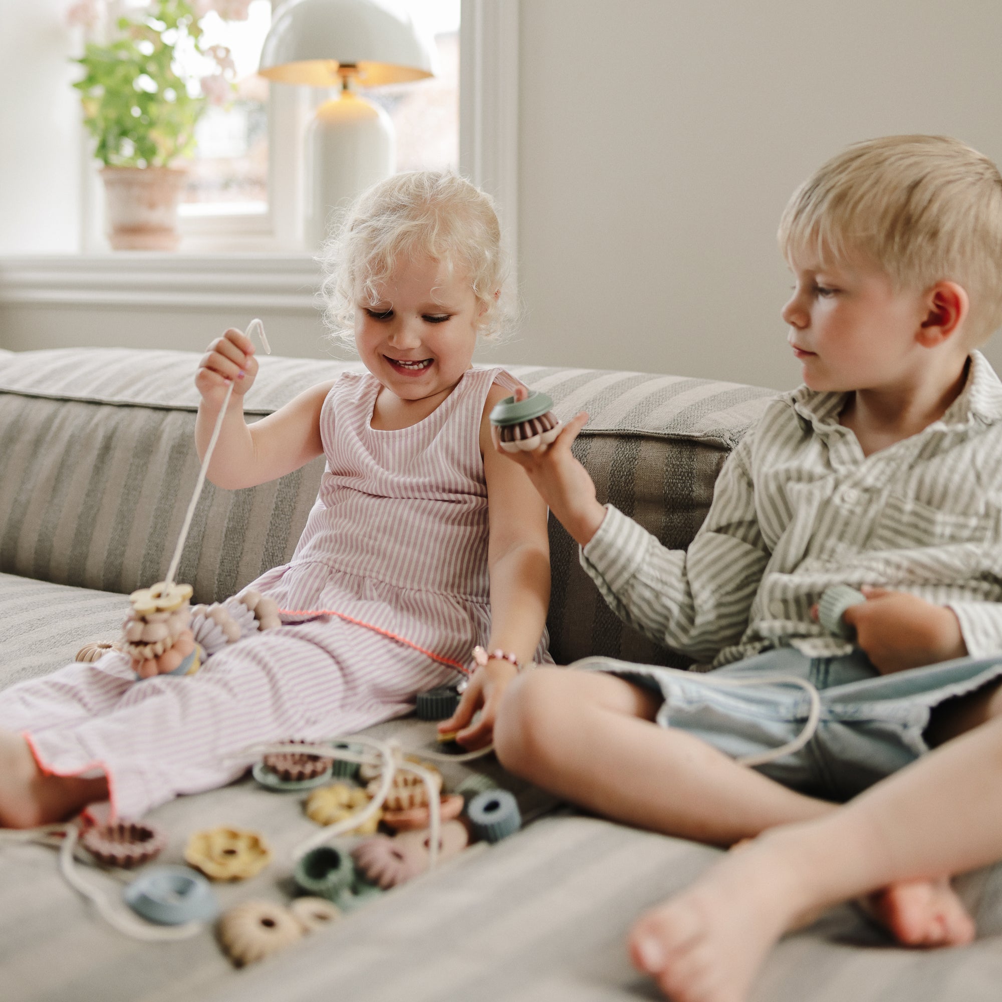 Two children use the mushie Bead Lacing Toy Set to build fine motor skills on a striped couch.