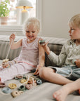 Two children use the mushie Bead Lacing Toy Set to build fine motor skills on a striped couch.