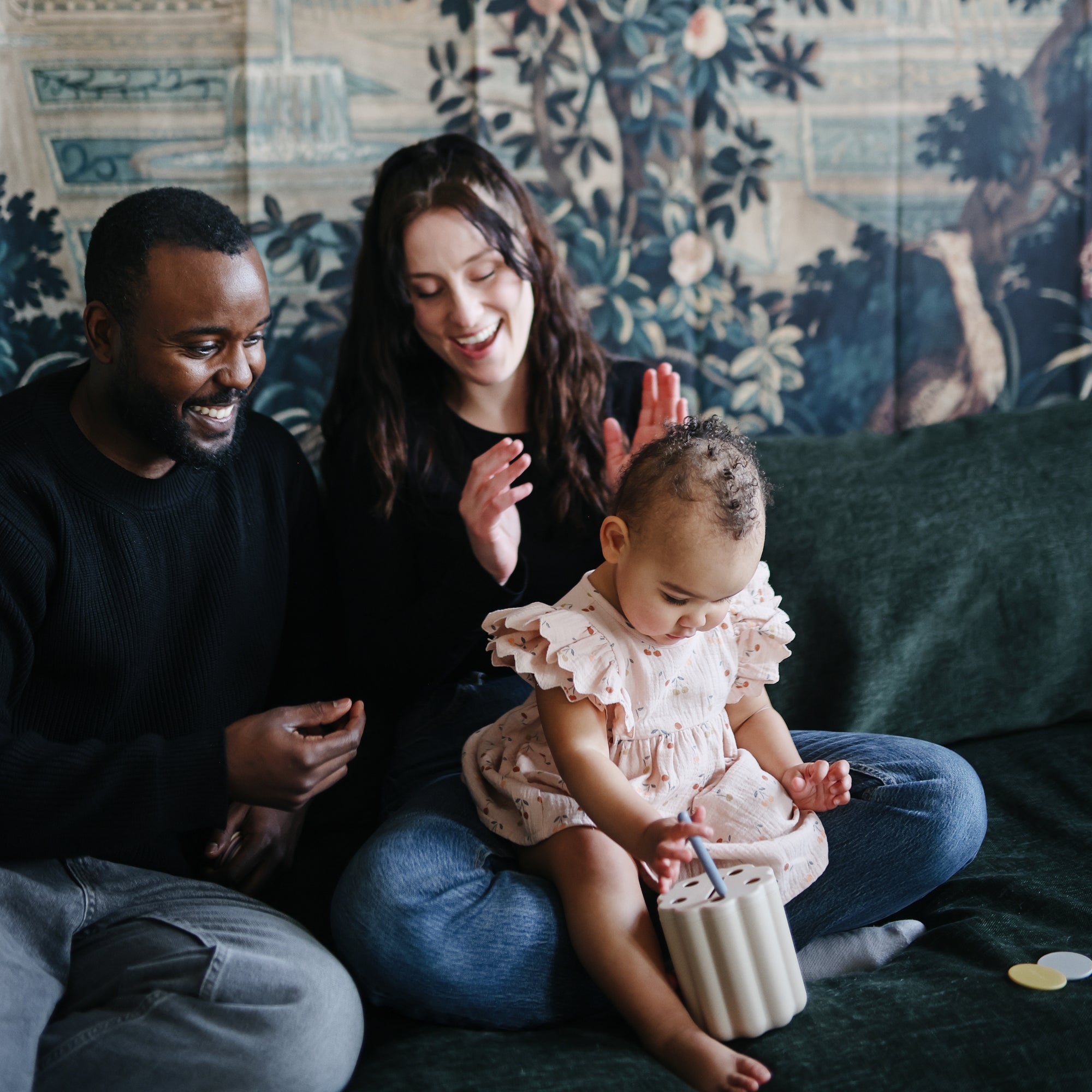 A smiling man and woman sit on a couch with a toddler girl in a pink dress, who is playing with the mushie Coin &amp; Tube Sorting Set. The adults look happy and engaged, and there’s a decorative tapestry in the background.