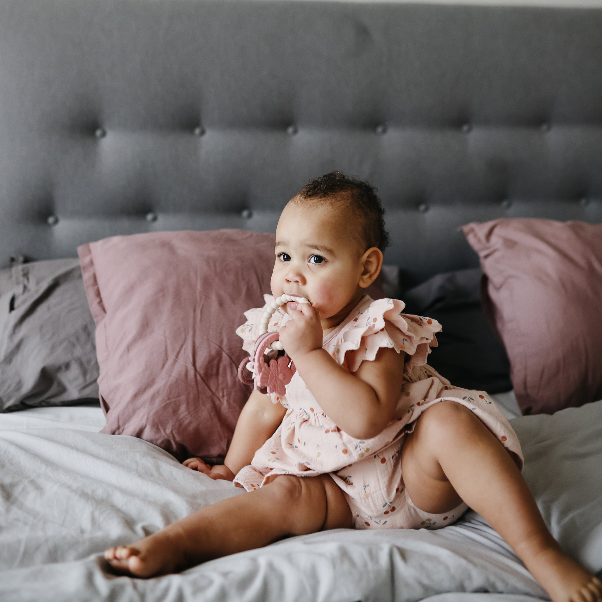 Baby in a pink dress chews the mushie Fairy Teething Ring on the bed.