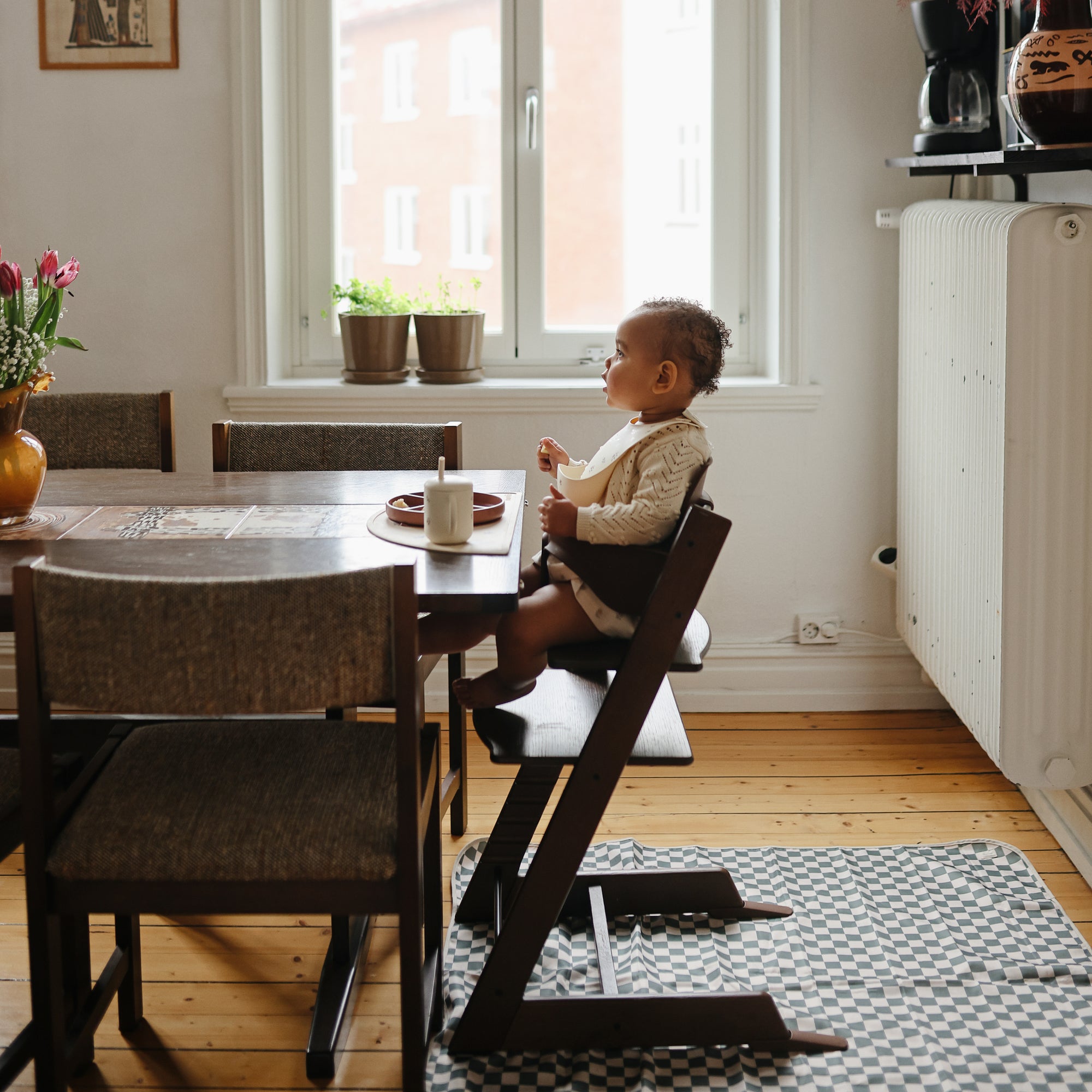 A young child sits in a high chair at the dining table, eating with a spoon. The Mushie Splat Mat protects the wooden floors, while potted plants on the windowsill soak up morning sunlight streaming through.