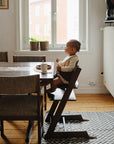 A young child sits in a high chair at the dining table, eating with a spoon. The Mushie Splat Mat protects the wooden floors, while potted plants on the windowsill soak up morning sunlight streaming through.