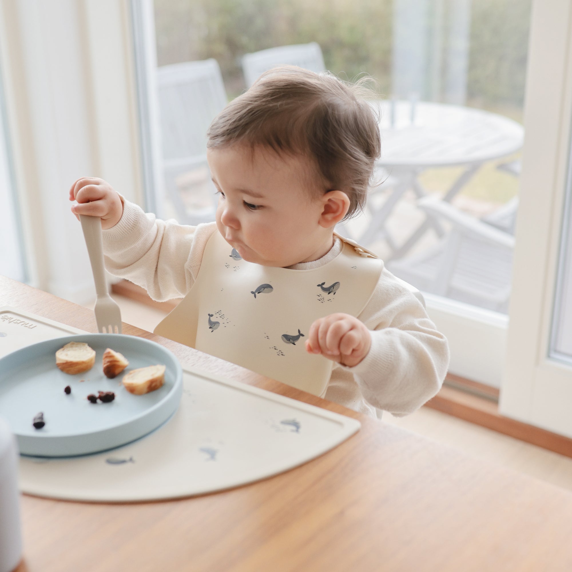 A toddler uses a mushie Silicone Placemat at the table with a whale-print bib.