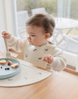 A toddler uses a mushie Silicone Placemat at the table with a whale-print bib.