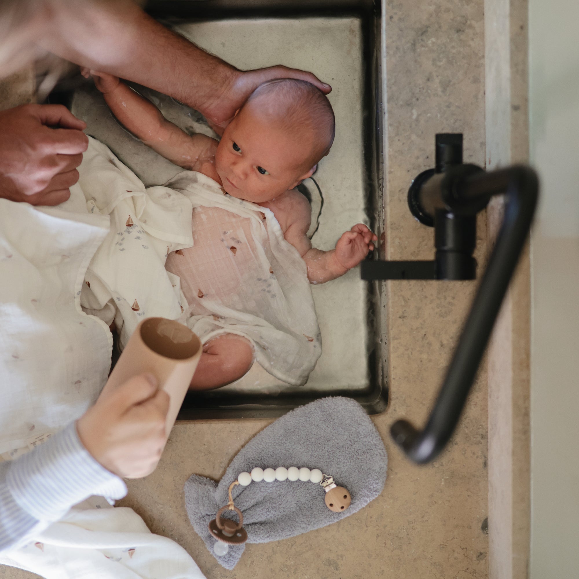 A baby wrapped in a light cloth is bathed in a kitchen sink with an adult’s support, while FRIGG Rope Silicone Pacifiers from FRIGG and a gray whale-shaped washcloth rest on the counter nearby.