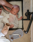 A baby wrapped in a light cloth is bathed in a kitchen sink with an adult’s support, while FRIGG Rope Silicone Pacifiers from FRIGG and a gray whale-shaped washcloth rest on the counter nearby.