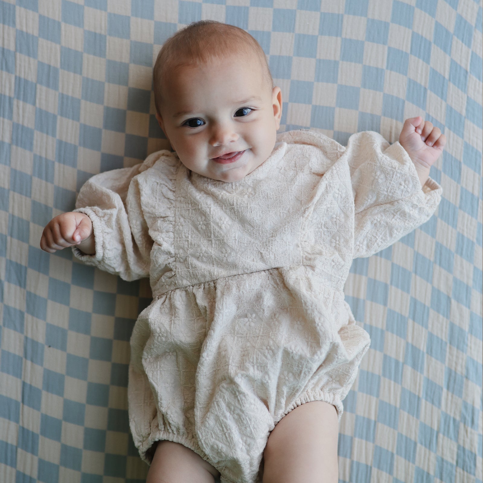 Smiling baby in a romper on mushie Extra Soft Muslin Crib Sheet with blue and white checks.