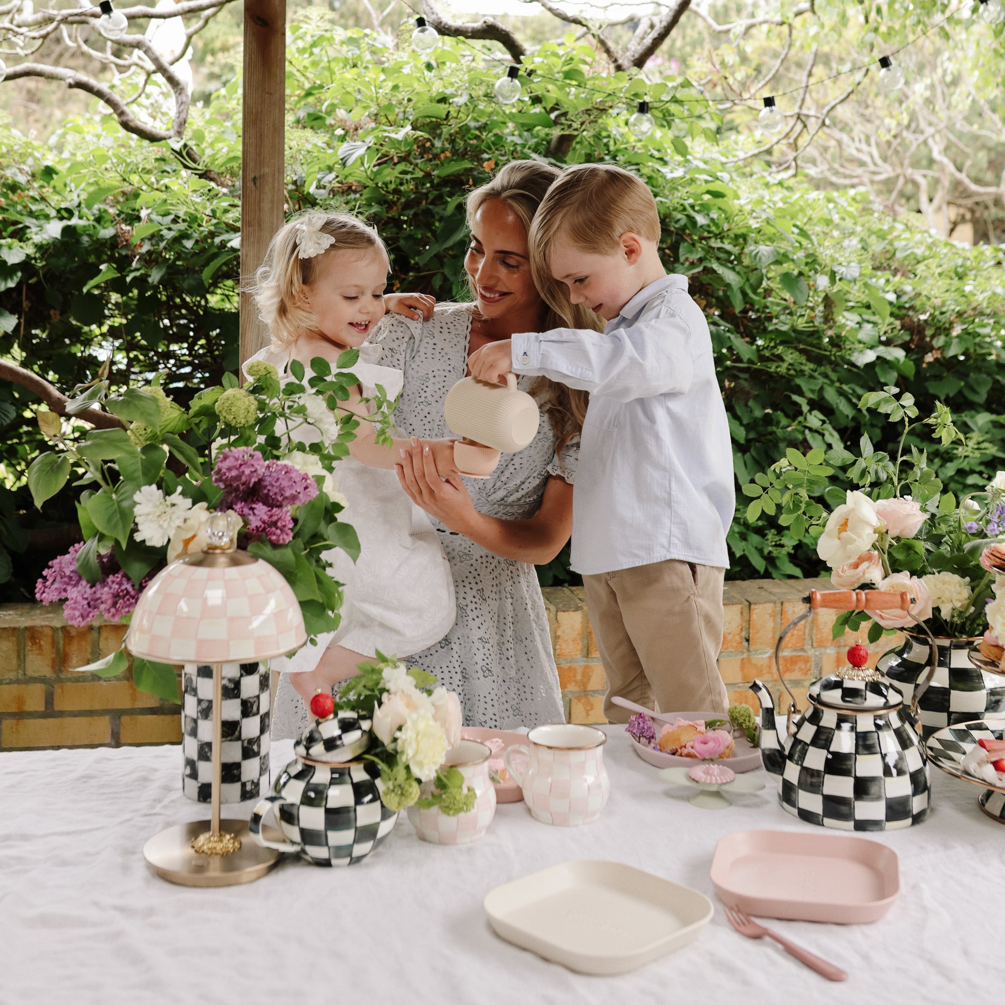 A woman and two kids use mushie's Dinnerware Fork and Spoon Set at a floral outdoor tea party.