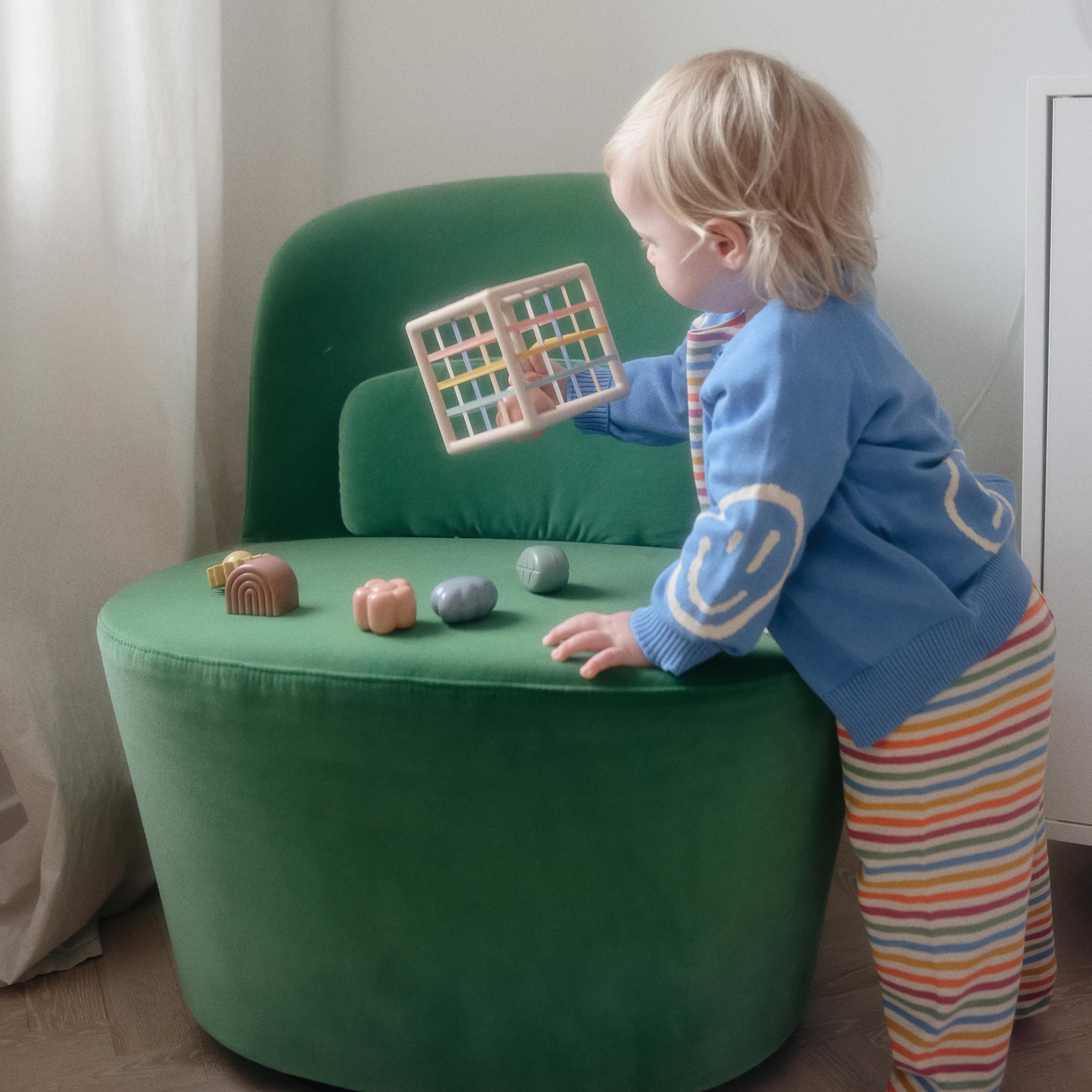 Toddler plays with mushie Elastic Shape Sorter, sitting in a green chair in striped pants.