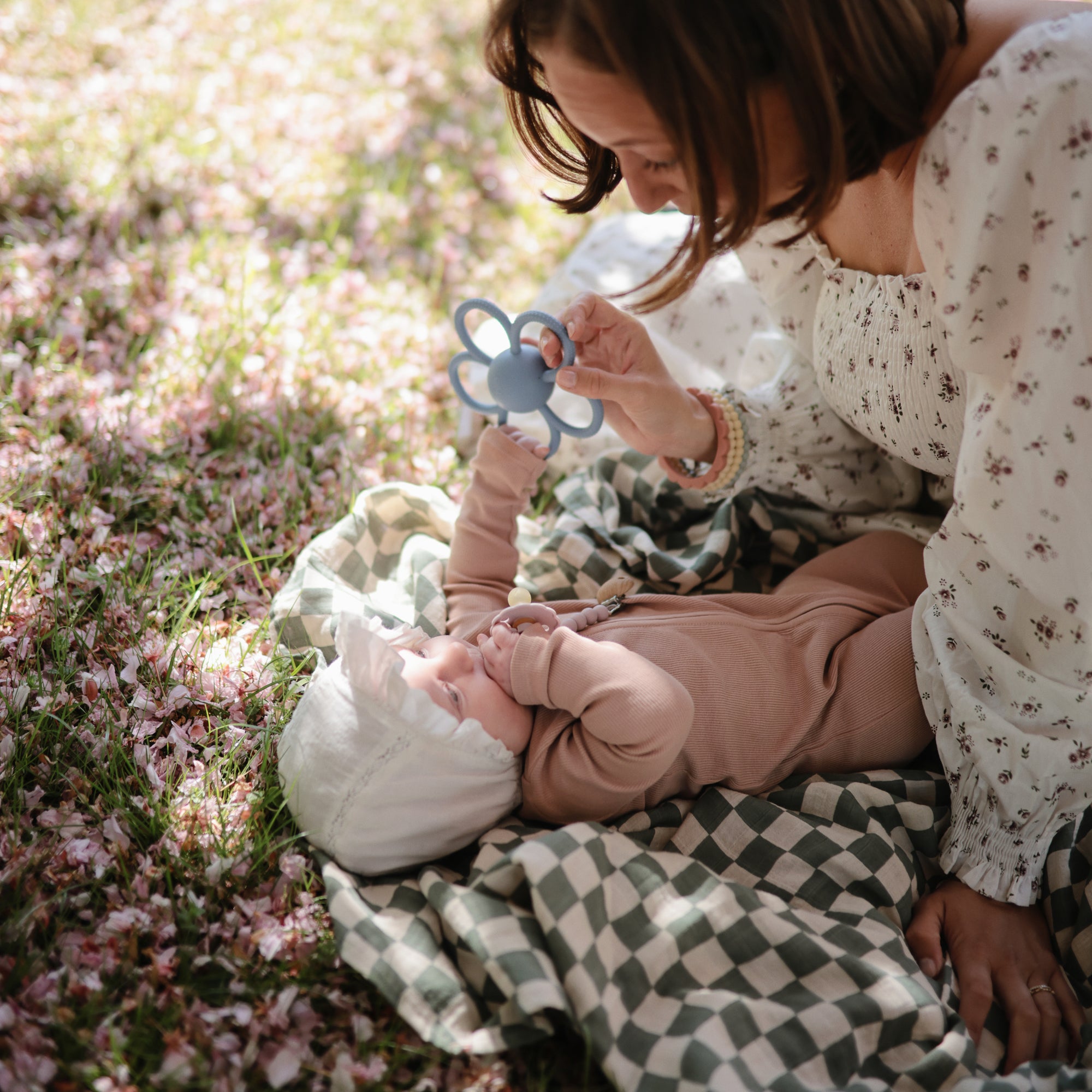 Woman and baby with mushie Daisy Rattle Teether among pink petals on a blanket outdoors.